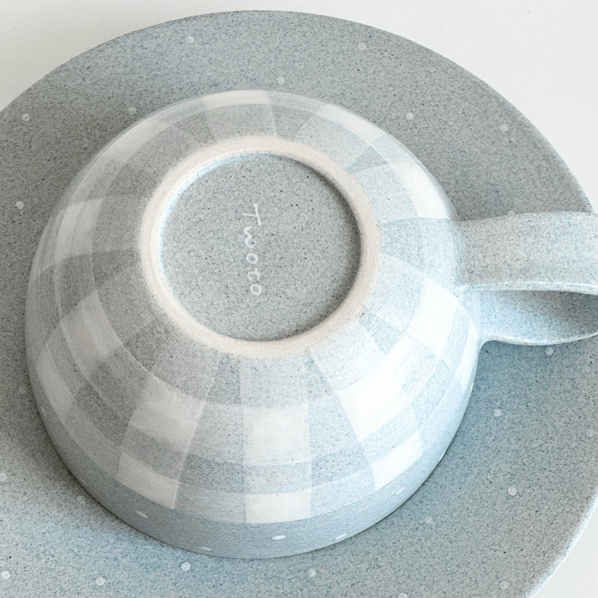 Bottom view of grey gingham ceramic cup on matching saucer, showing Twoto maker's signature